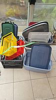 Photo showing multiple large plastic totes in blue and gray, several lids, a black collapsible crate, and yellow, red, green plastic baskets stacked together.