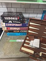 Stack of vintage board game boxes including Sudoku 2, Tri-Ominos, Conspiracy, Stratego, Conflict War Game, and Stock Ticker on countertop.