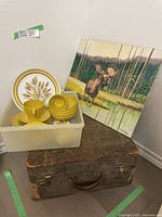 Photo showing worn vintage suitcase, Melmac dishes with wheat design, and original moose painting