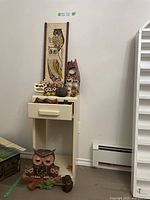 Overall lot showing vintage owl figurines displayed on and around a small side table, plus a wooden panel with painted owls mounted behind.