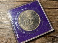 Close-up of the reverse side of the coin showing the crowned initials "EP" and commemorative inscriptions on a purple background inside protective case.