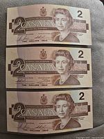 Front view of three Canadian two-dollar bills aligned vertically, each showing Queen Elizabeth II portrait and denomination details.