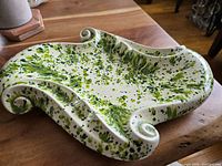 Top and angled view showing the full ashtray with green and yellow splatter glaze on an off-white base, curled corner details, and overall design