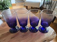 Four indigo ombre footed cocktail glasses shown from side angle on wooden table with plant in background.