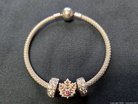 Front view of the Pandora sterling silver bracelet displaying the Canadian maple leaf charm with central red gemstone, flanked by two rhinestone clips on a mesh band with round engraved clasp.