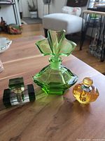 Three vintage colored glass perfume bottles displayed on a wooden surface. One large green bottle with flared stopper, one small dark green and clear bottle, and one small yellow bottle with rounded stopper and silver collar.