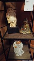 Photo showing all three Asian statues on shelving unit: standing Laughing Buddha on black base, sitting Buddha in colorful paint, and white elder figure with staff.