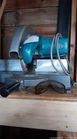 Side profile image of a blue-green Makita chop saw on a grey metal stand, showing the blade guard and handle.