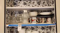 Shelf with clear glass pitcher, multiple clear drinking glasses, white bowls and plates, coffee cups with floral design, and black baking trays.