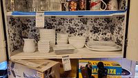 Shelves showing multiple white plates stacked, bowls of different sizes, small condiment dishes, and a ceramic pitcher. Some plates have a floral design.