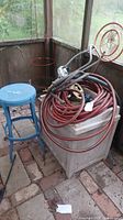 Photo showing red garden hose coiled on a white plastic chest, blue painted wooden stool, and copper-colored metal sprinkler frame near a screened porch corner.