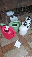 Photo showing four watering cans in different colors (red, green, white) and two white weed sprayers with black handles arranged on a brick floor.