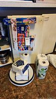 Shows a bundle of packaged kitchen paper goods such as plastic wrap, storage bags, cooking liners in rack holder above and below, plus rolls of paper towels, wet wipes, and napkins on counter with plastic lazy Susan trays.