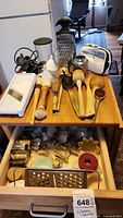 Kitchen utensils and appliances arranged on a wooden surface and inside an open drawer, showing various items including cookie cutters, graters, wooden spoons, and a hand mixer.
