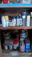 Shelf with multiple spray paint cans including yellow Rust-Oleum, HD brand cans, and brown colored stains or finishes. Lower shelf has Olympic brand paint cans and various smaller jars and bottles.