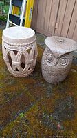 Two ceramic plant stands shown together on outdoor mossy ground against a wooden wall background, one owl-shaped and one cylindrical with arches