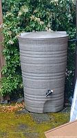 Full view of the rain barrel standing outdoors on a paved area with greenery in the background. Shows textured ribs and the tap. Barrel is topped with a fitted lid.
