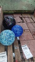 Three decorative glass gazing balls sitting on wooden planks; one dark blue with colorful mosaic, one cobalt blue mottled, and one light blue spotted.