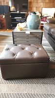 Photo showing brown tufted storage ottoman in front of a grey slatted modern coffee table, with decorative items on table.
