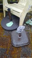 Photo showing two umbrella stands with bases on outdoor ground beneath plastic chair, highlighting condition and material.