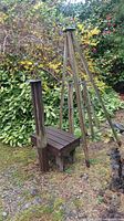 Wooden bench, tall backrest visible, wooden tripod, and metal sundial on wooden base in outdoor garden setting surrounded by foliage.