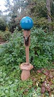 Wide angle photo showing the wooden post with blue gazing ball on top, ceramic mushroom at the bottom, and gargoyle figure in the middle, surrounded by green landscaping