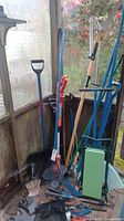 Photo showing assortment of yard tools including shovels, rakes, pole saw, and green gardening seat leaning against a wall inside a shed.