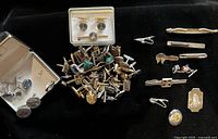 Overview of metal case with cufflinks, loose pile of assorted cufflinks and tie bars, and row of additional tie clips