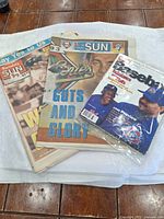 Photo showing three baseball-related magazine and annual covers spread on a white cloth on a wooden table. Two Sunday Sun magazines and one white-covered baseball magazine with player photos.