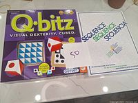 Q-bitz board game box showing title, puzzle cards, and dice cubes with numbered dots and colored sides, along with a paper tag marked '50'.