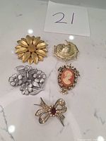 Five assorted brooches arranged on a white marble surface: gold-tone sunflower, silver-tone floral with hummingbird, textured silver-tone swirl, gold-tone cameo with red background, and gold-tone bow with red jewel.