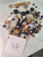 Photo showing a large pile of assorted buttons in multiple colors and sizes laid on a white surface with a ruler for scale and a note with '42'.