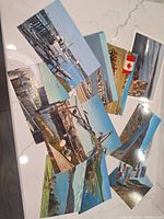 Photo showing a spread of multiple colorful unused Canadian postcards with coastal scenes including boats, docks, lighthouses, and rural landscapes on a white marble surface.