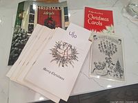 Overview photo showing multiple vintage Christmas ephemera items on a white surface including Christmas Ideals booklet, Christmas Carols booklet, blank Christmas cards, and a Division Detectives greetings booklet.