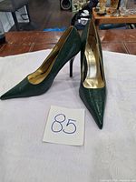 Pair of dark green women's high heel pumps, pointed toe, slim stiletto heel, gold interior lining with branding visible on insole.