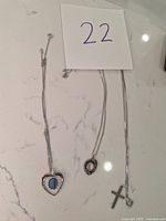 Three silver necklaces laid on a white marble surface, showing pendants and chains with a paper note marked '22'.