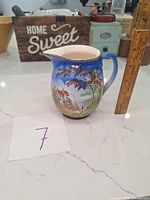 Front and side views of the ceramic cream jug showing painted scenic cottage and waterfront, ruler for scale indicating approx 5 inches tall.
