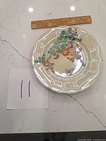 Top view of two vintage china plates with hollyhocks floral design, butterflies and insects, placed side by side with a ruler above for scale.