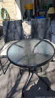 Photo of a round tempered glass outdoor patio table with black metal legs accompanied by two black wicker patio chairs with armrests and beige cushions.