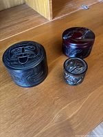 Three different carved lidded boxes shown together on floor: black Haida-style stone box with large tribal face, reddish-brown stone box with horse carving, smaller dark wooden carved box