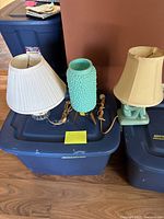 Three small table lamps on a blue storage bin: left is the sitting girl teal lamp, center is the teal bubble lamp on wooden legs, right is the beige shade lamp on teal base.