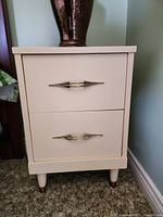 Light-colored bedside table with two drawers and metallic handles against a wall.