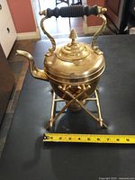Front view of brass teapot with wooden handle, spout, hinged lid, set on brass stand with measuring tape showing height