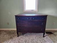 Front view of solid wood chest of drawers showing four drawers with two small and two large drawers, metal ring pull handles, on carpeted floor against a light green wall.