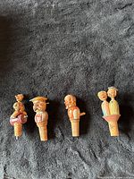 Top view of all four hand carved cork stoppers showing detailed figures including a drummer, man with mustache, bearded man, and two figures in a basket.
