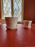 Two off-white McCoy pottery planters with matching saucers, displayed on a red surface with a window in the background.