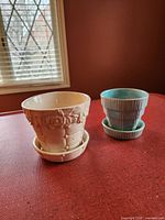 Photo showing both McCoy pottery planters and their matching saucers on a red surface near a window with blinds.