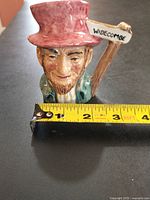 Miniature Toby mug with red top hat and sign saying 'Widecombe' next to measuring tape showing approx. 3 inches height.