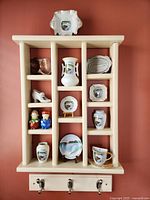 Photo of the shadowbox containing 12 Niagara Falls themed ceramic souvenirs. The shadowbox is cream colored and divided into nine compartments with three metal hooks at the bottom.