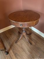 Full view of the round wooden table showing the turned pedestal base with three curved legs, drawer with circular metal pull, and the table's finish.